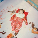 chinese baby laying on bed wrapped in red flower swaddling clothes with red pillow and red dot on forehead
