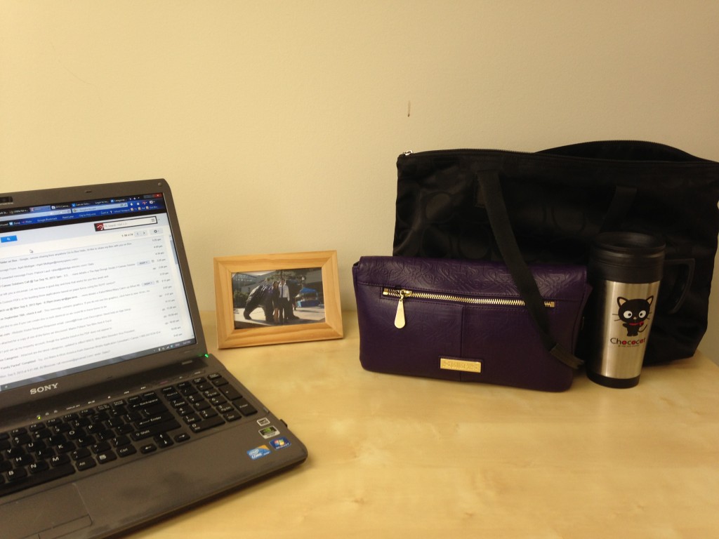 light wood desk with laptop, wooden framed picture, purple purse, black bag, and chococat mug