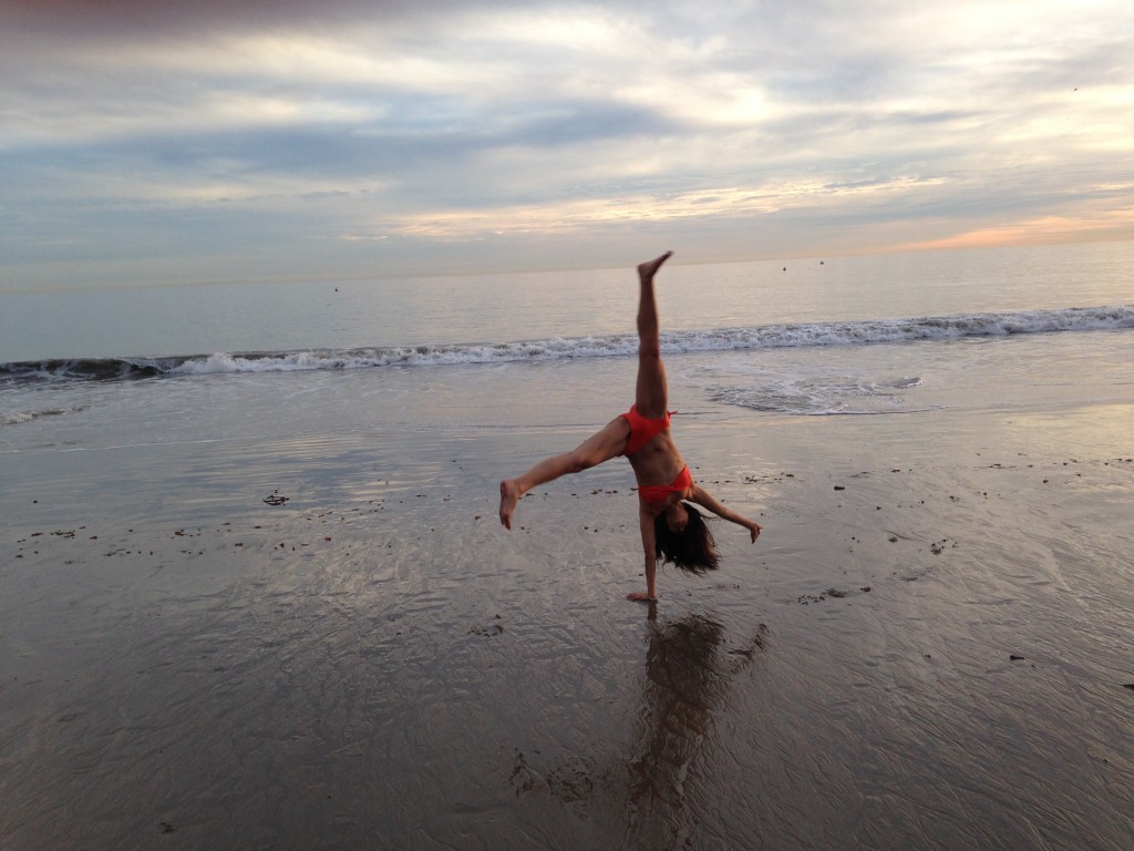 girl in orange bikini doing cartwheel at beach paused on one hand