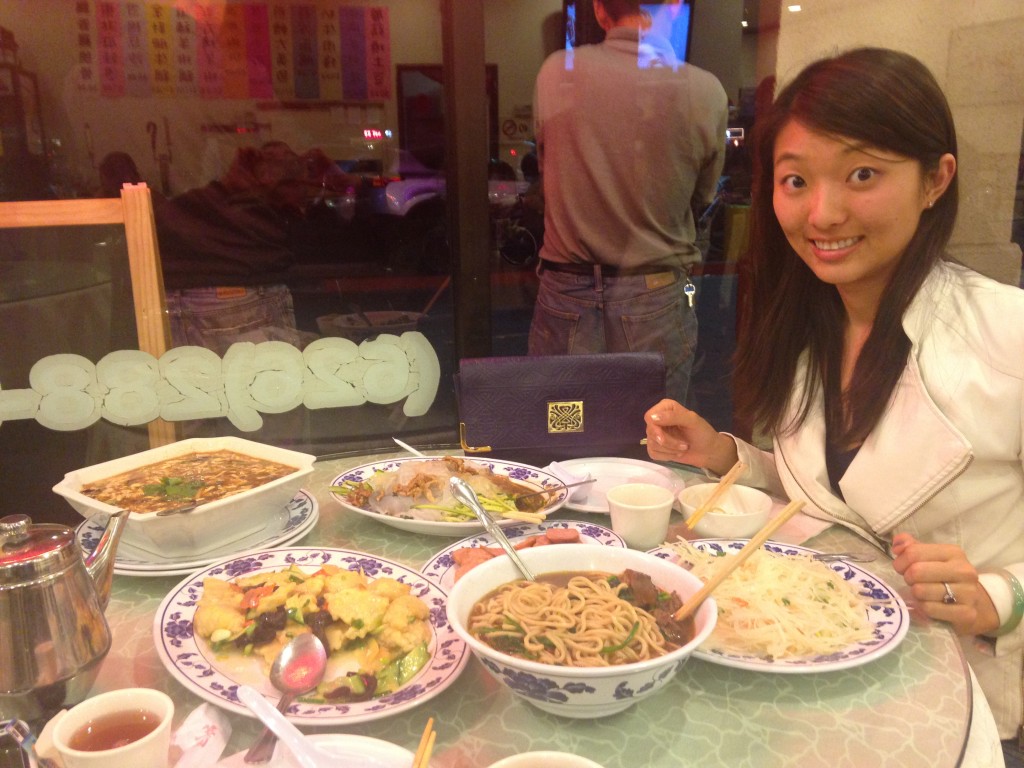 table filled with giant dishes of northern chinese food and girl looking skeptical about two people eating it all